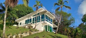 Une maison moderne à deux étages, dotée de grandes fenêtres et d’un toit plat, entourée d’une végétation luxuriante et de palmiers.