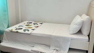 A small, single-bed room with a white bedspread and pillows, a window with blinds, and a curtain.