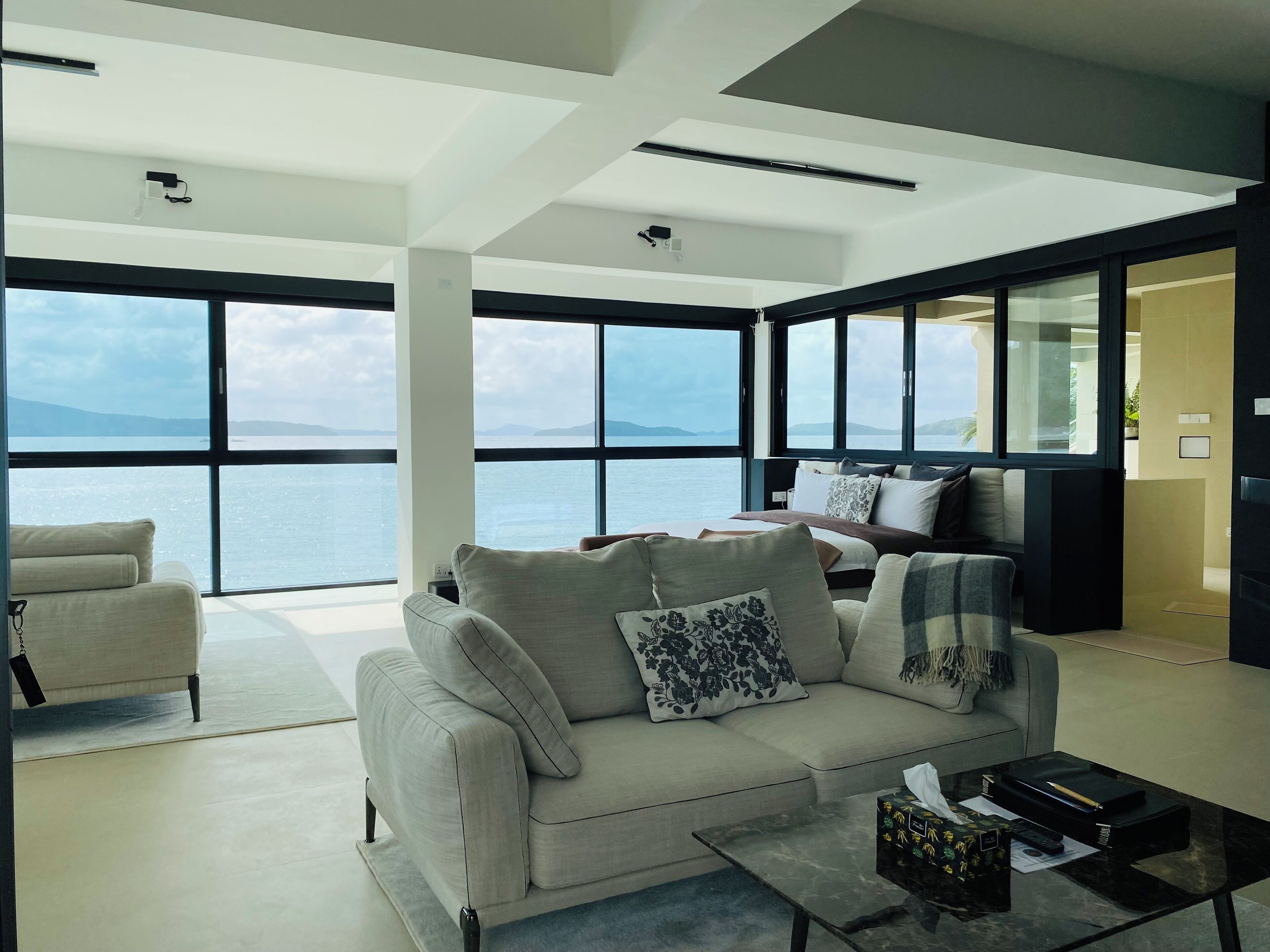 A modern living room with a large window, a sofa, a coffee table, and a bed with a view of the sea.
