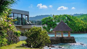 A modern building with a glass balcony overlooking a body of water, a thatched structure on a rock, and lush greenery in the background. - The Waterfront Studio (Busuanga)