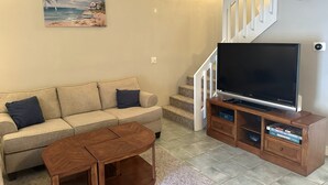 A living room with a beige sofa, a coffee table, a TV on a wooden stand, and a painting of a beach scene. - The Blue Turtle: 3-bedroom, 2-bath MB townhouse w/ 5 min walk to the beach (Myrtle Beach)
