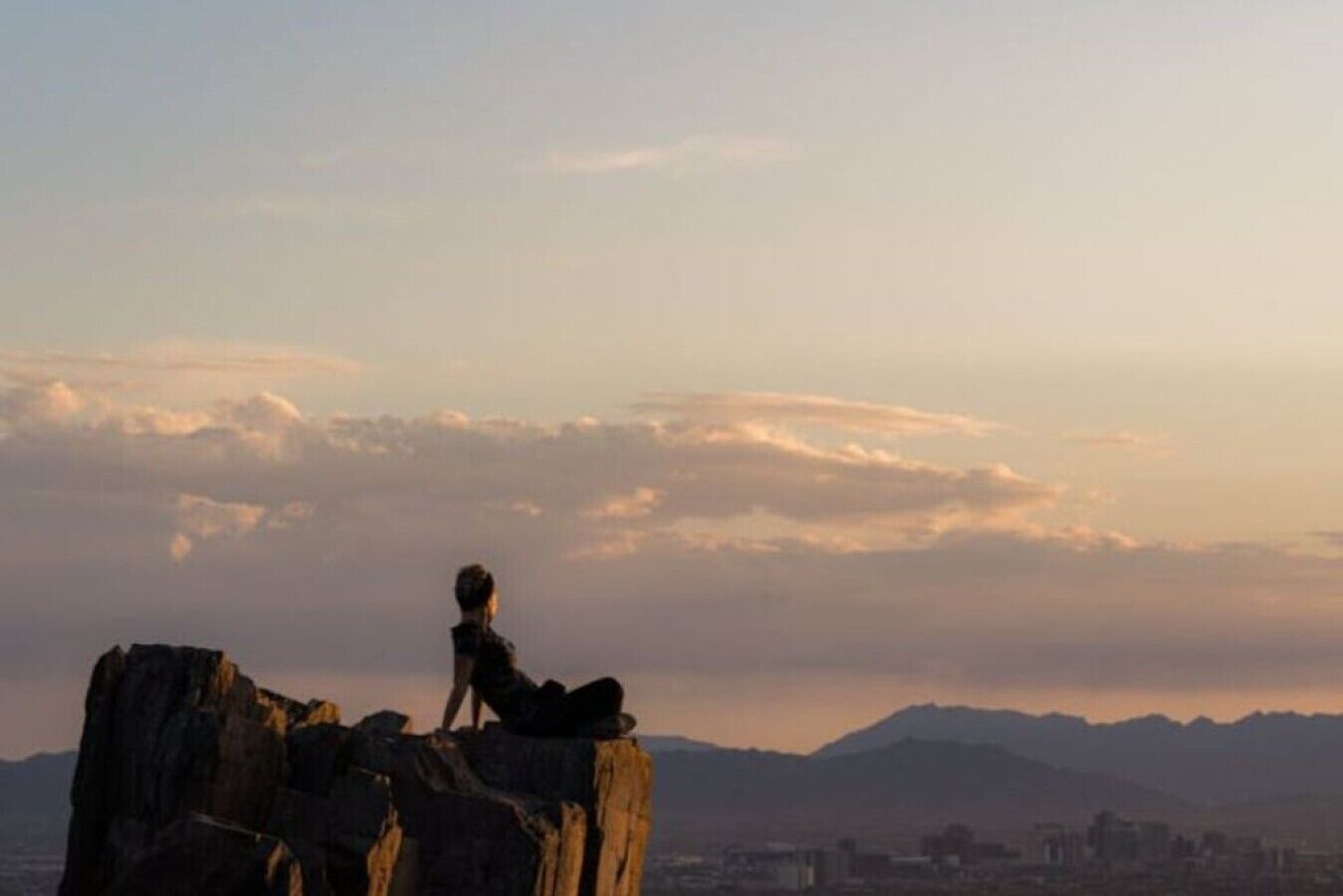 A person standing on a rock formation with a cityscape and mountains in the background.