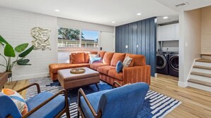A modern living room with a sectional sofa, a coffee table, and a blue armchair. There is a large window with a view of trees and a white fence. A laundry area with a washing machine and dryer is visible in the background. - Old Town Scottsdale Condo | Pool | Patio | Grill (Scottsdale)