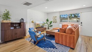 A modern living room with a sofa, chairs, a coffee table, and a TV. There is a wooden cabinet with glassware and a plant. The room has white walls and a blue rug. - Old Town Scottsdale Condo | Pool | Patio | Grill (Scottsdale)