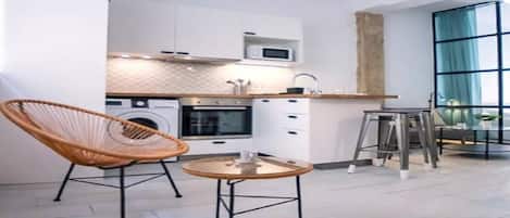 Una cocina moderna con armarios blancos, microondas, horno y un lavamanos. Se ve una silla de mimbre y una mesita redonda con una taza.