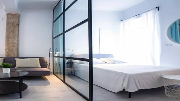 Et moderne hotelværelse med glasvæg, seng, sofa, lille rundt bord og en potteplante.