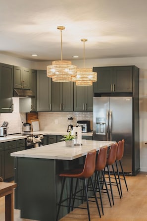 A modern kitchen with dark green cabinets, a large island with bar stools, stainless steel appliances, and a unique woven light fixture.