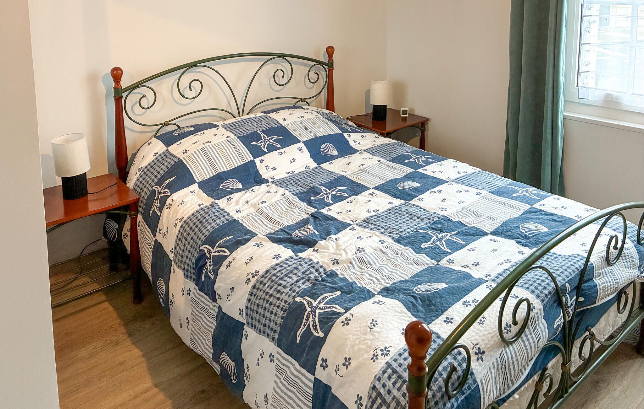 Un lit avec une couette bleue et blanche, une tête de lit en bois, deux tables de chevet avec des lampes et une fenêtre avec des rideaux.