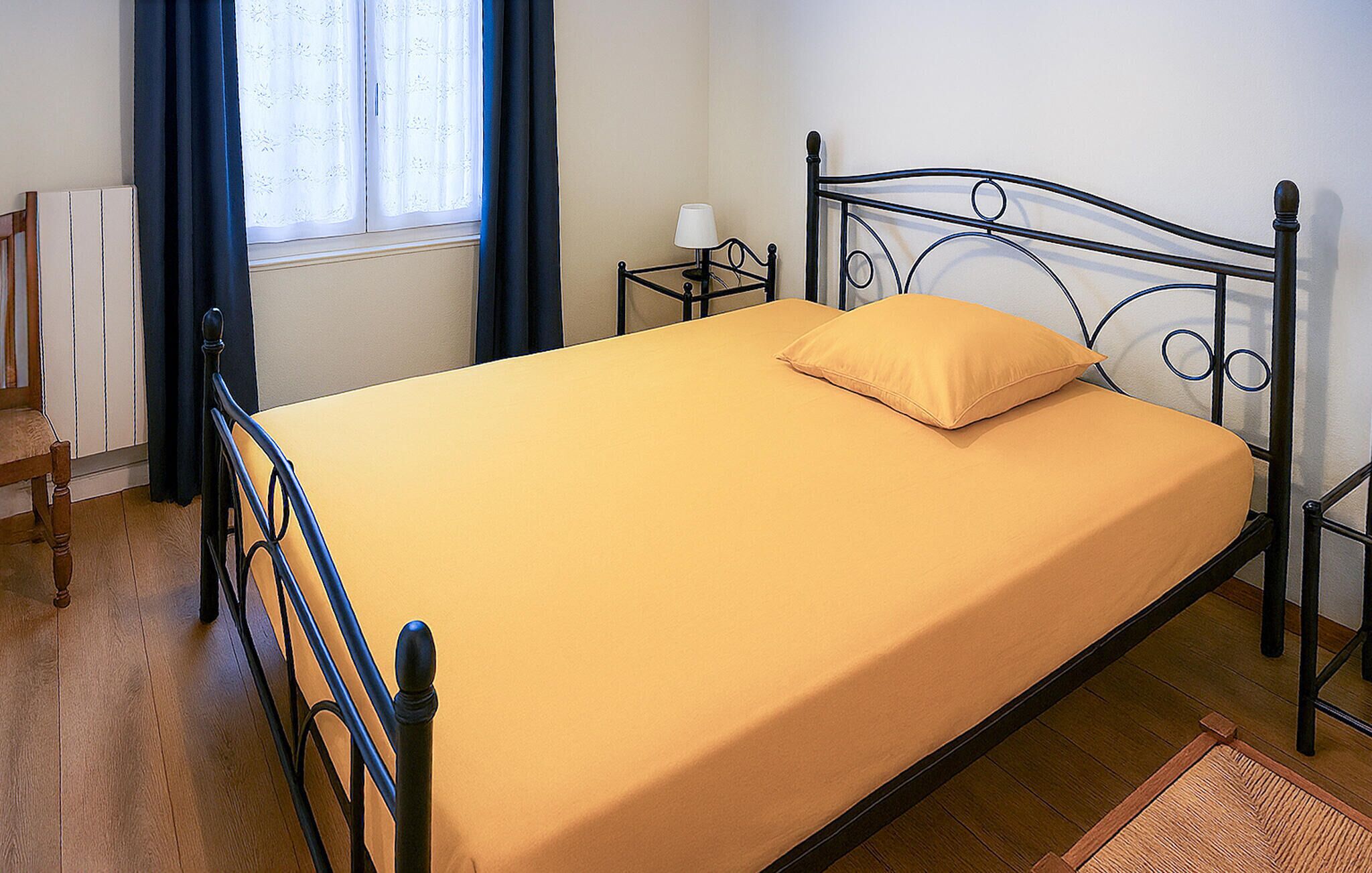 Ein Einzelbett mit gelber Matratze und passendem Kopfkissen in einem Zimmer mit Holzboden, einem Fenster mit Vorhängen und einem Nachttisch mit Lampe.