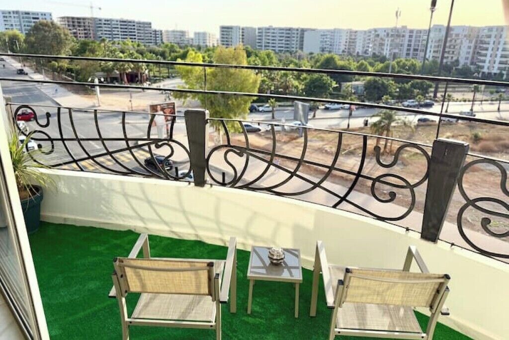 A balcony with a view of a cityscape, featuring a railing, two chairs, a small table, and a potted plant.