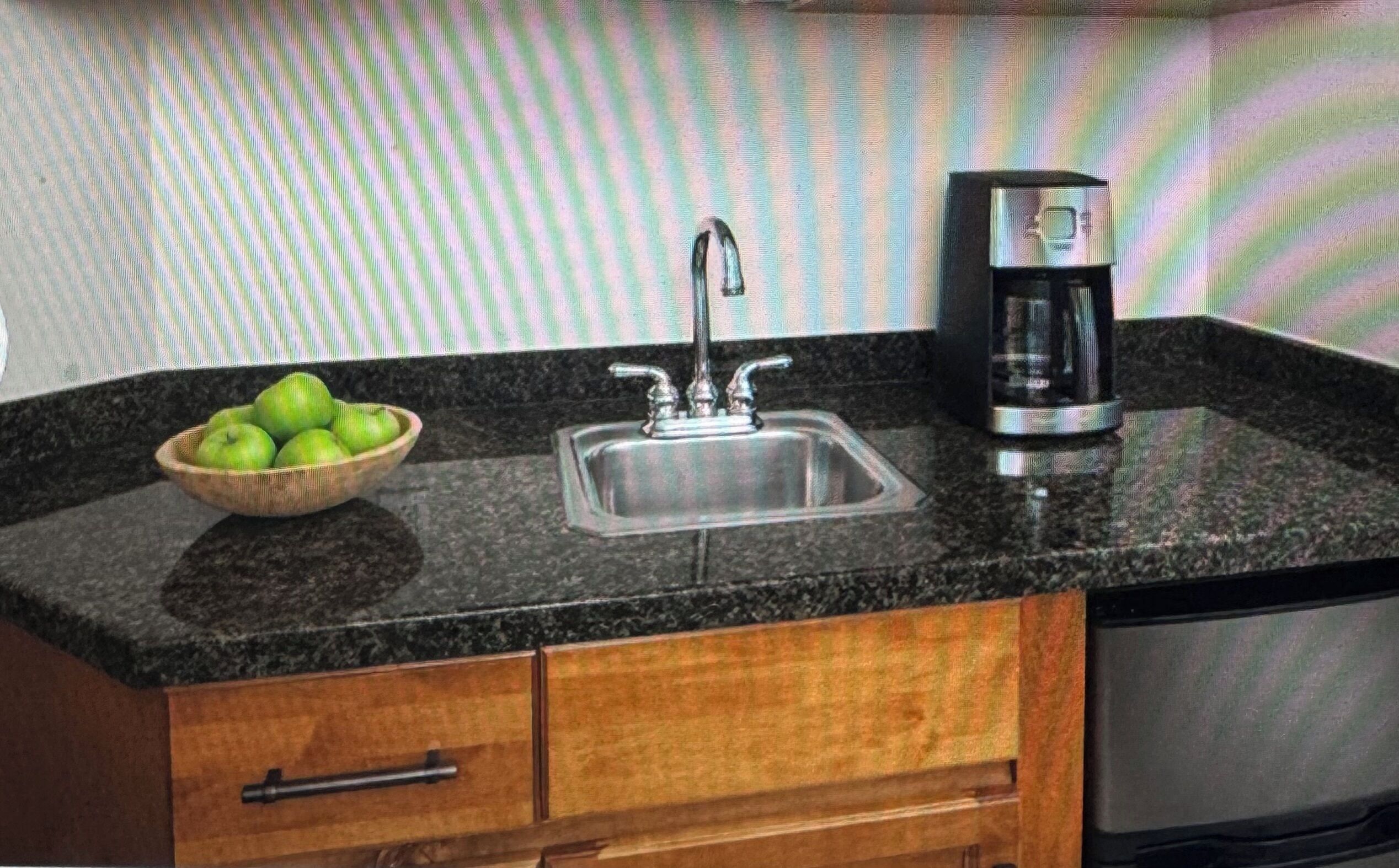 A kitchen with a granite countertop, a stainless steel sink, a coffee maker, and a wooden cabinet with a bowl of green apples.