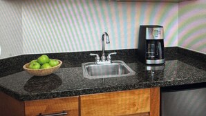 Una cucina con un piano di lavoro in granito, un lavello in acciaio inossidabile, una macchina da caffè e un mobiletto in legno con una ciotola di mele verdi.