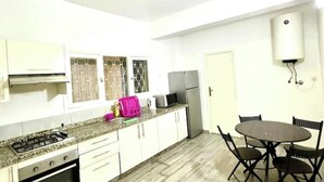 A compact kitchen with white cabinets, a gas stove, a microwave, and a refrigerator. There is a dining table with four chairs and a water heater mounted on the wall. - AR Apparts (city center -Wifi 100 Mb) (Fès)