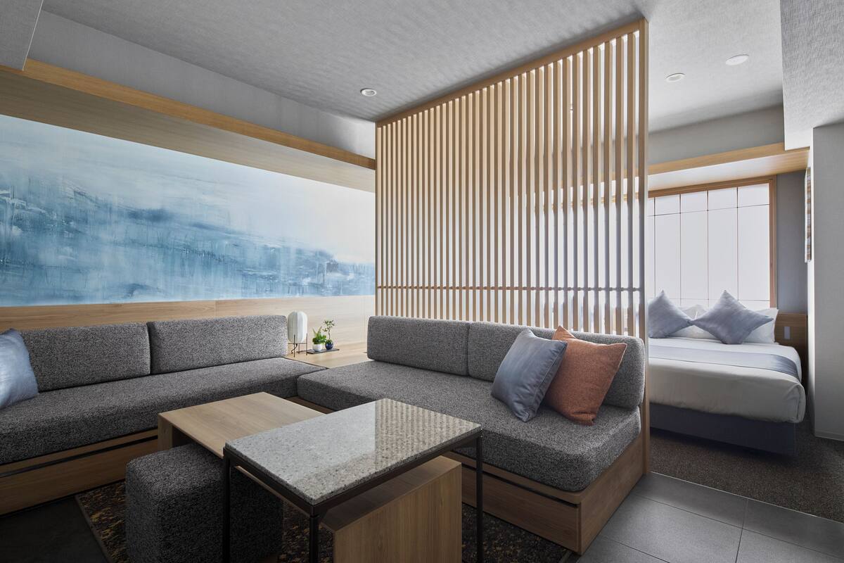 A modern hotel room with a sectional sofa, a small table, a bed with pillows, and a large abstract painting on the wall.
