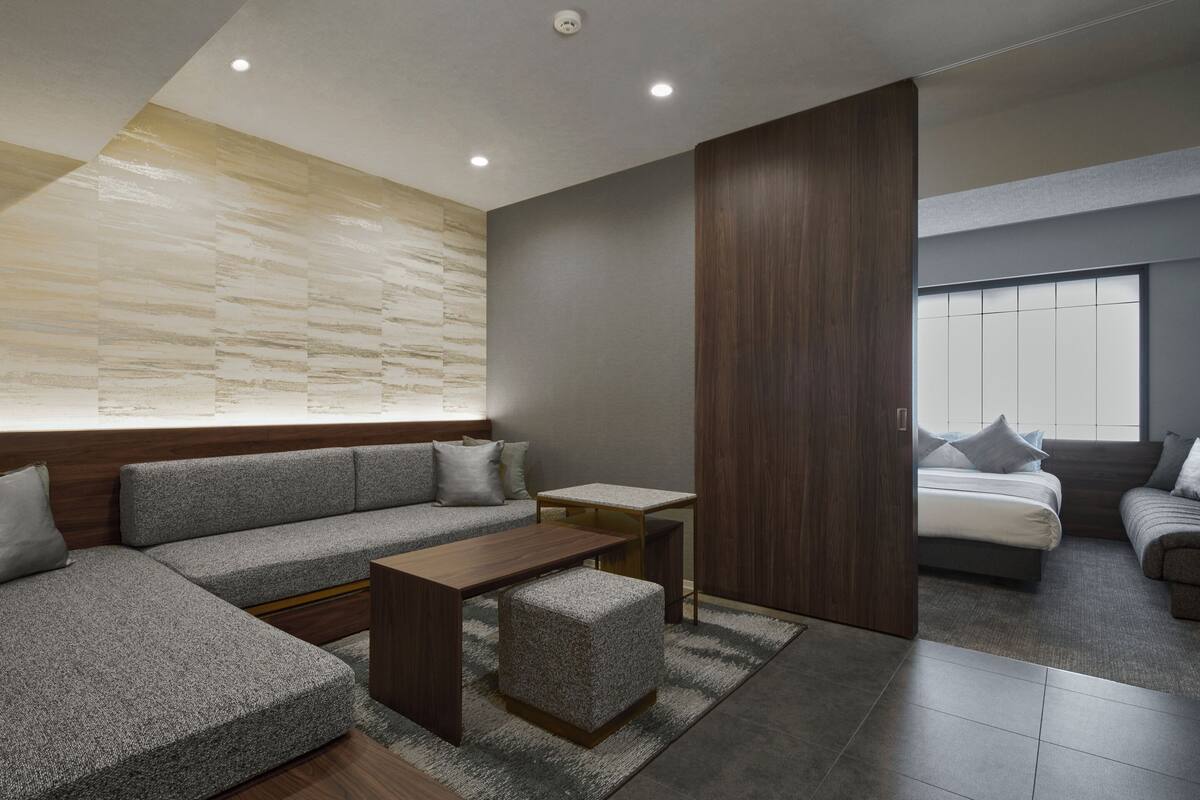 A modern living room with a sectional sofa, a coffee table, and a small ottoman. There is a wooden accent wall and a large window with blinds.