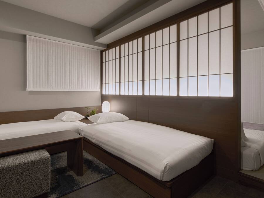 A hotel room with two beds, a desk, and a large window with blinds.