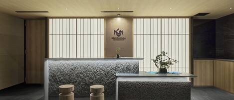 A reception desk with a logo, a potted plant, and a small table. There are two wooden stools and a textured bench.