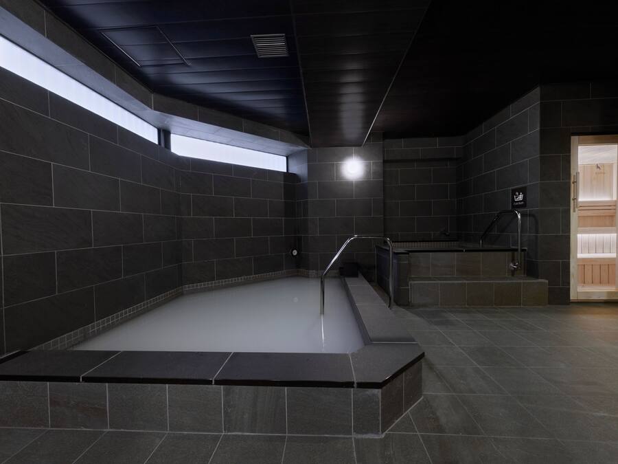 Un área de spa moderna con una bañera de hidromasaje rectangular, paredes de azulejos oscuros y un tragaluz.