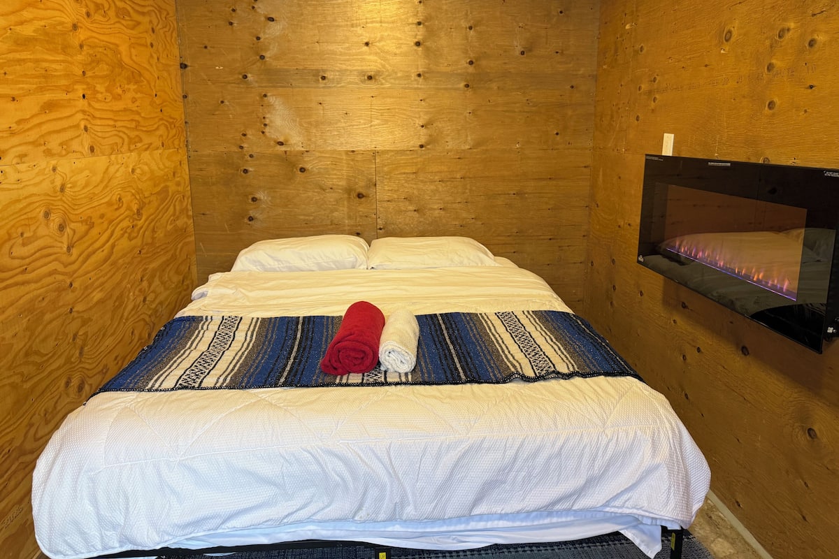 Um quarto pequeno e acolhedor com parede de madeira, cama com roupa de cama branca, toalha vermelha e televisão na parede.