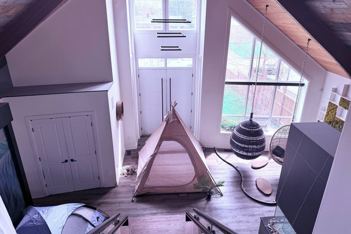 A loft-style room with a wooden floor, a tent-like structure, a hanging chair, and a large window.