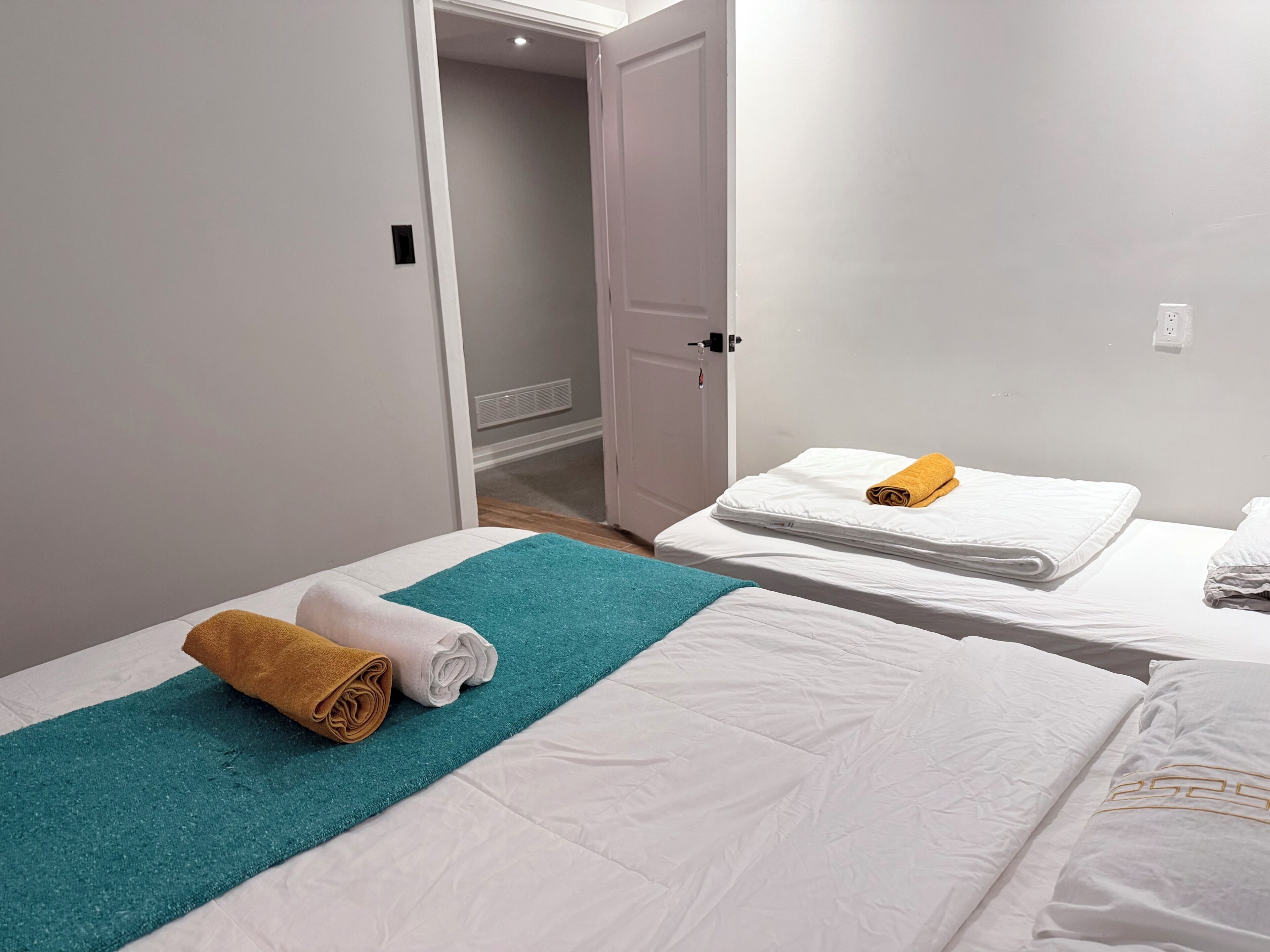 A hotel room with two beds, each with a white mattress and a teal throw blanket. There are towels on the beds, and a door leading to another room.