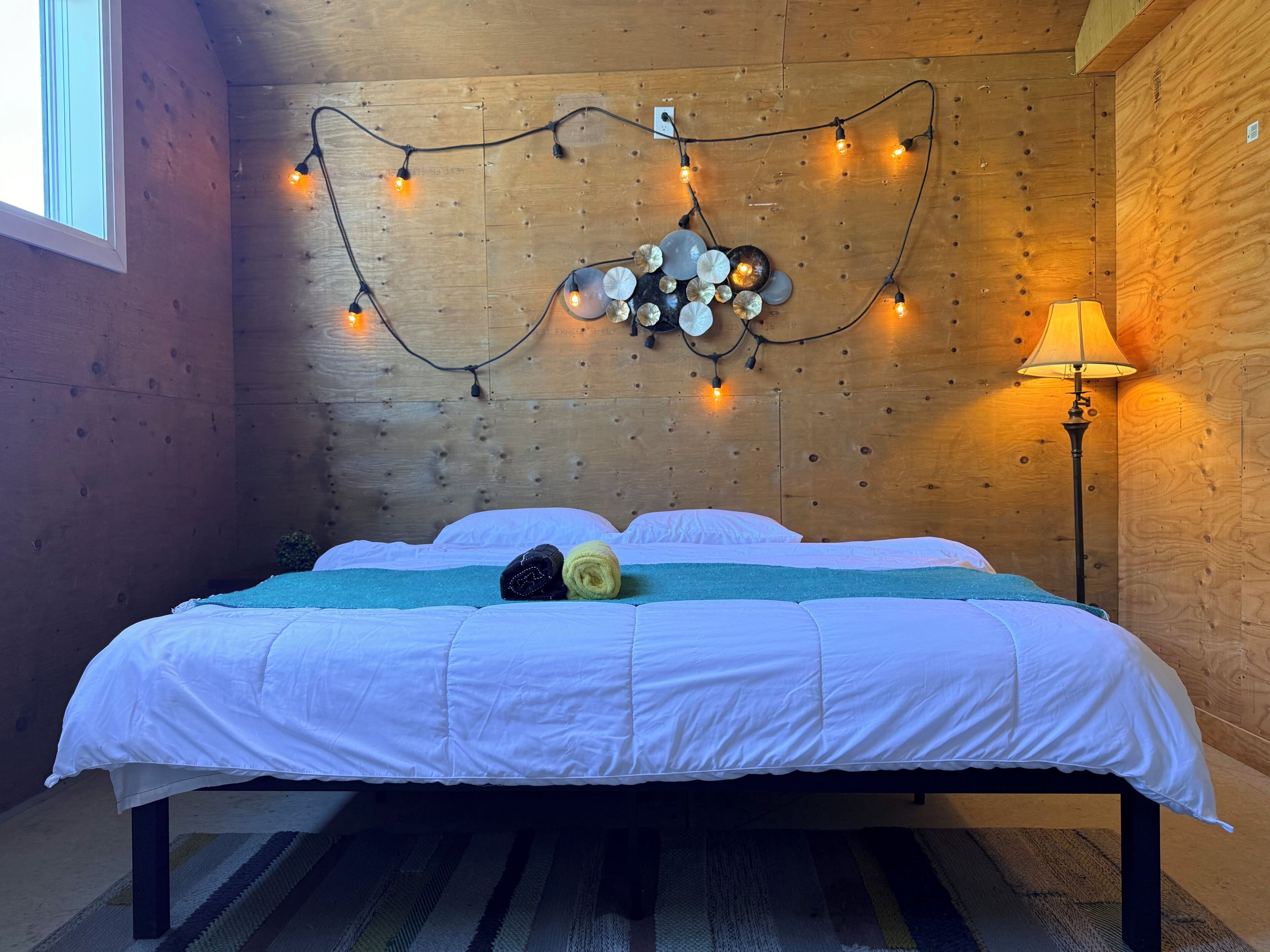 A wooden-paneled room with a bed, string lights, and a lamp.