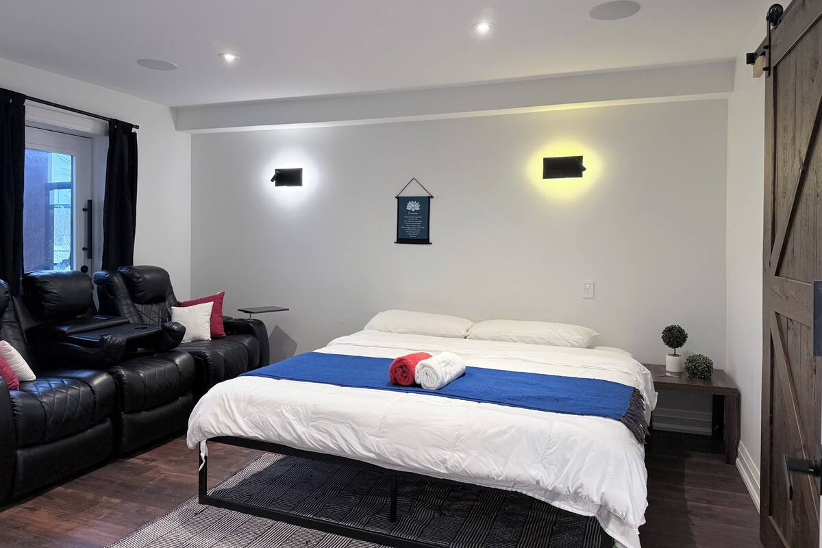 A modern bedroom with a bed, a leather sofa, a small table, and a wall-mounted light fixture.