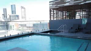 Une piscine sur le toit offrant une vue sur la ville, un espace salon couvert et une rambarde en verre.