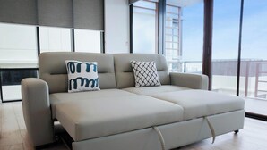 Un canapé moderne doté d’une section escamotable, de deux coussins décoratifs et offrant une vue sur le paysage urbain grâce à de grandes fenêtres.
