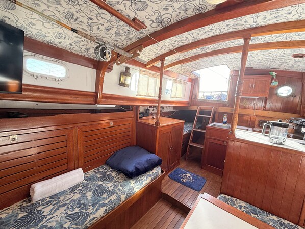 A compact cabin with wooden walls and a patterned ceiling, featuring a bed with a blue pillow, a small table, and a window.