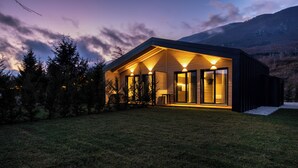 A modern house with a sloped roof, large glass windows, and a well-lit porch area. - The Village Chalet (Castel di Sangro)