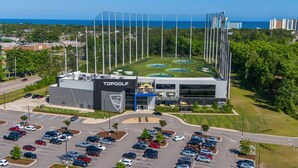 En TopGolf golfbane med stor parkeringsplads og en moderne bygning med udsigt over golfbanen.