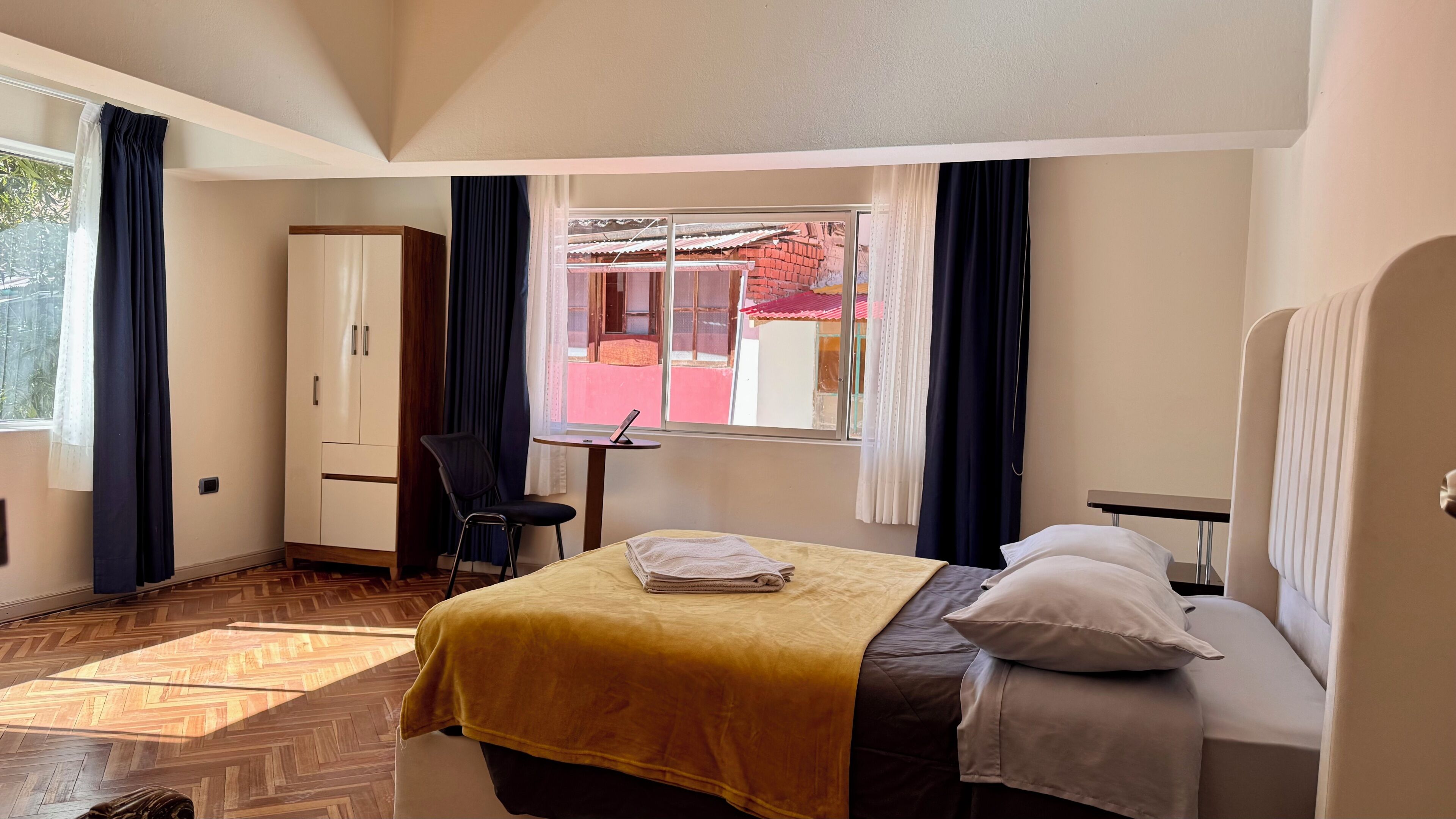 Une chambre d’hôtel comprenant un lit, un bureau, une chaise, une armoire et une fenêtre donnant sur les bâtiments environnants.