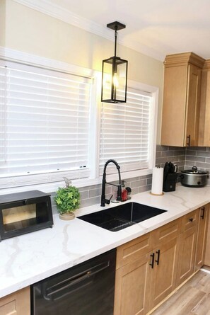 A kitchen with a double sink, wooden cabinets, a black microwave, and a hanging light fixture. - Welcome to University Place (Ruston)