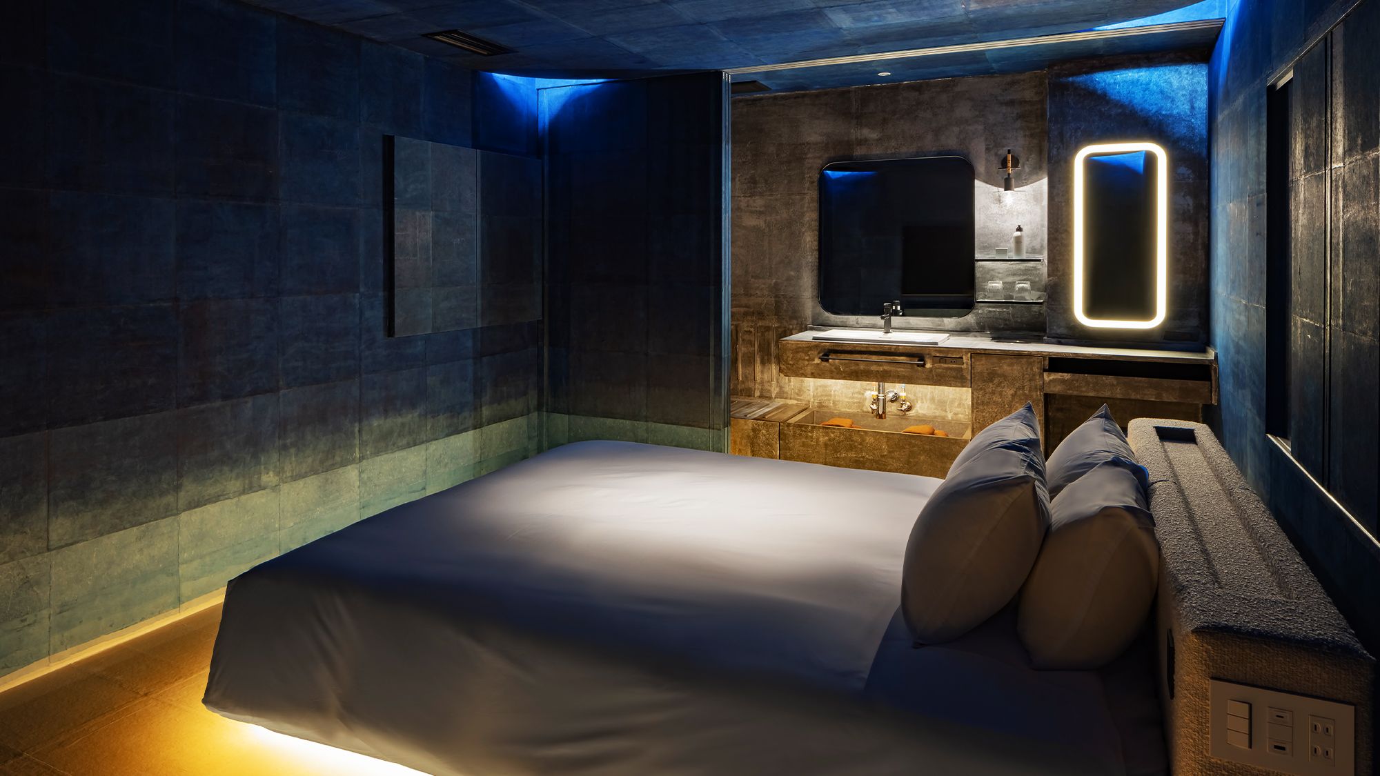 A modern bedroom with a large bed, a built-in bench with storage, a mirror, and a small bathroom area with a sink and mirror.