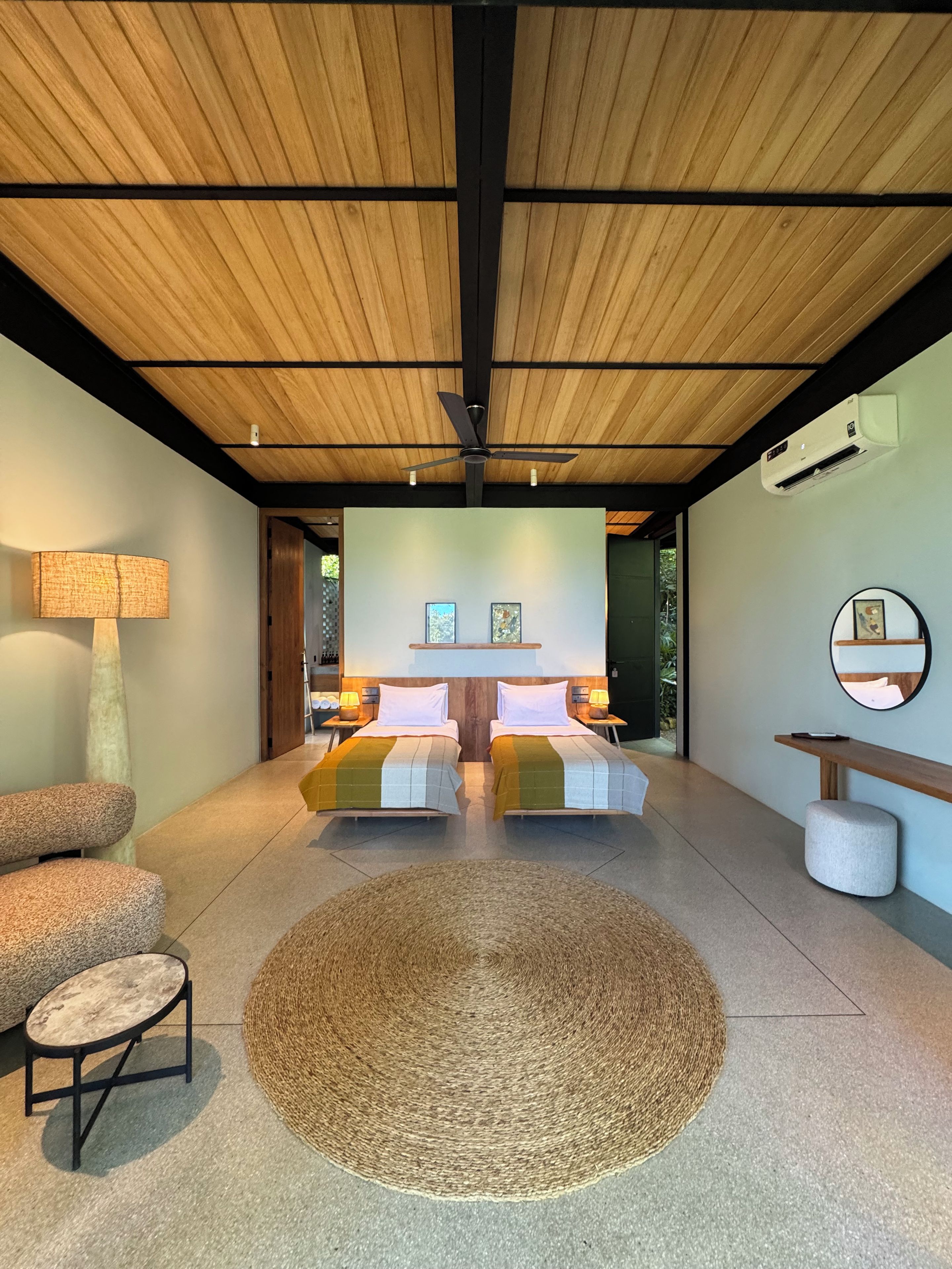 Un dormitorio moderno con techo de madera, dos camas, un tapete redondo tejido y una mesita de noche con lámpara.