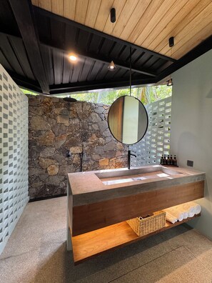 A modern bathroom with a stone wall, a wooden ceiling, a large mirror, and a built-in sink. - The Sevana (Matara)