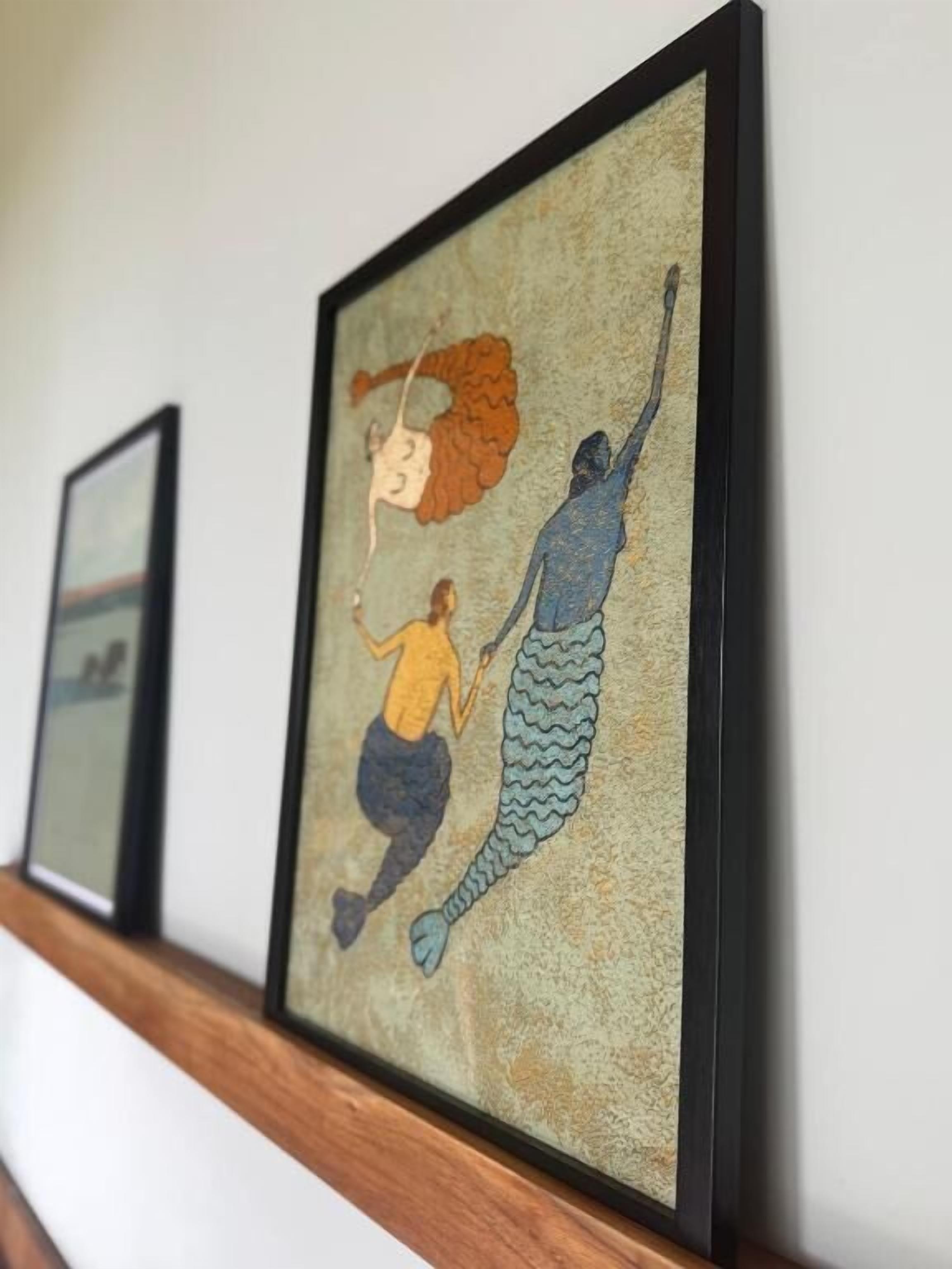 A framed artwork of two figures with tails on a wall.
