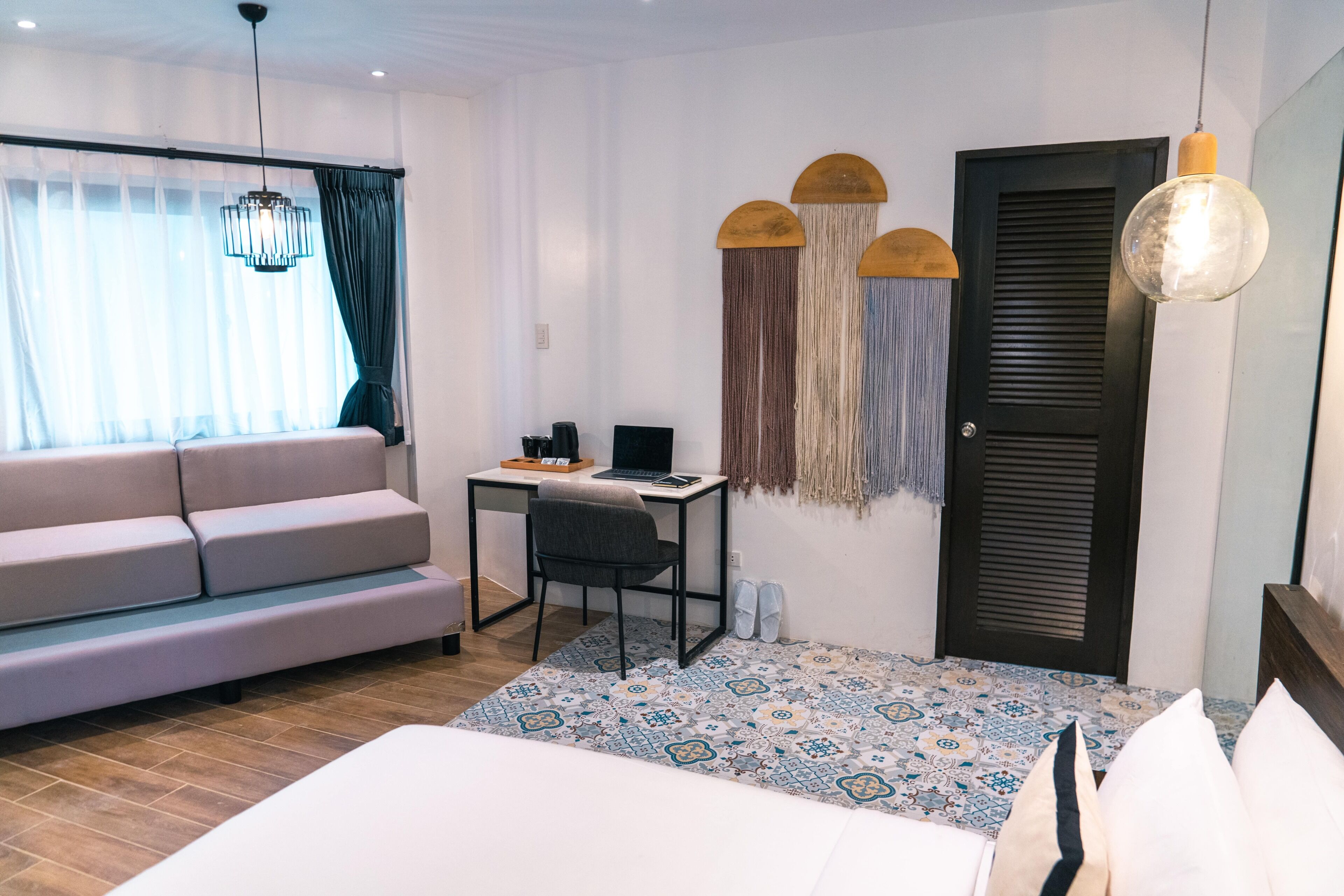A modern hotel room with a bed, a desk, a sofa, and a large window with curtains.