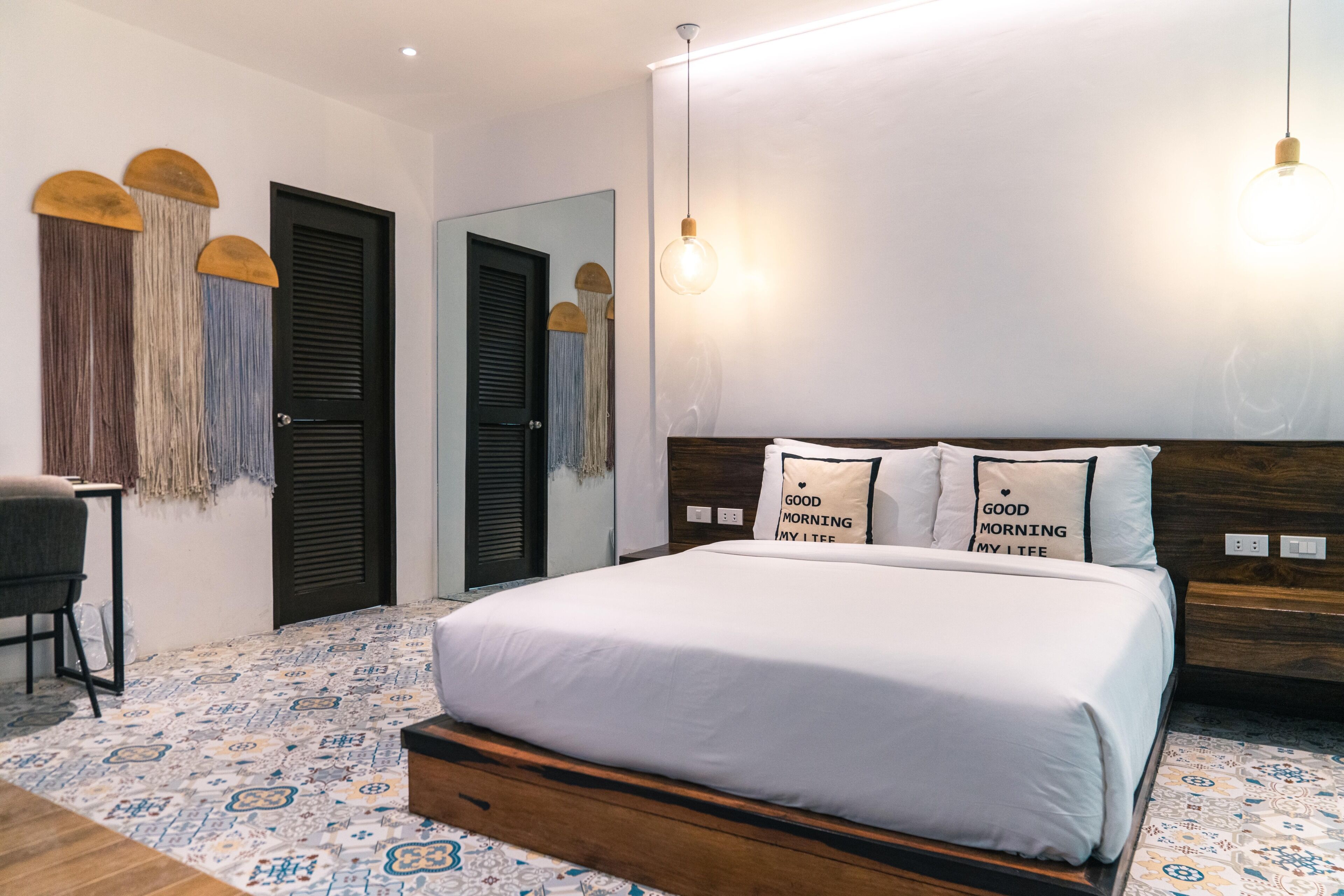Un dormitorio con cama, dos puertas, una silla, lámpara colgante y suelo estampado.