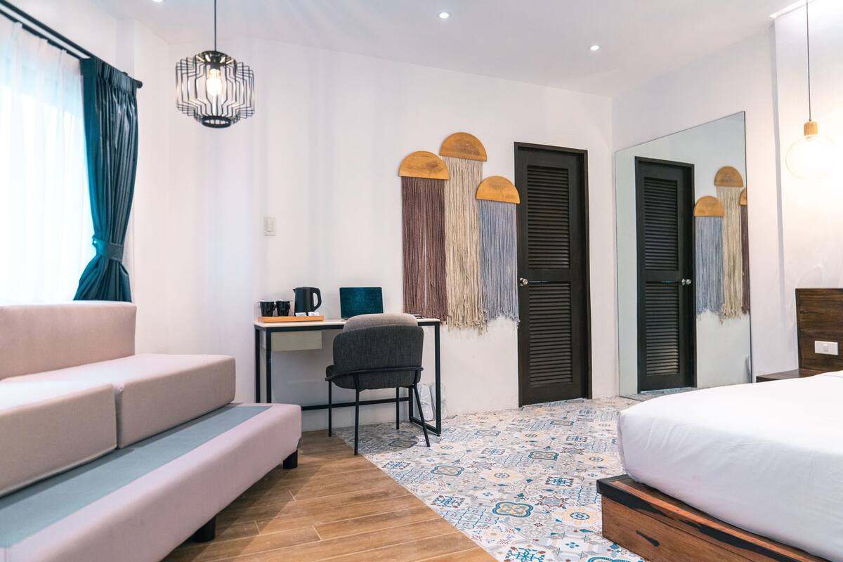 A bedroom with a bed, a desk with a chair, a sofa, a hanging light fixture, and a mirror.