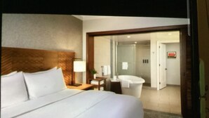 A hotel room with a large bed, a bedside table with a lamp, a bathroom with a bathtub, and a door leading to another room. - Beautiful full resort access -2-bedroom condo with AC in delightful Lahaina (Lahaina)