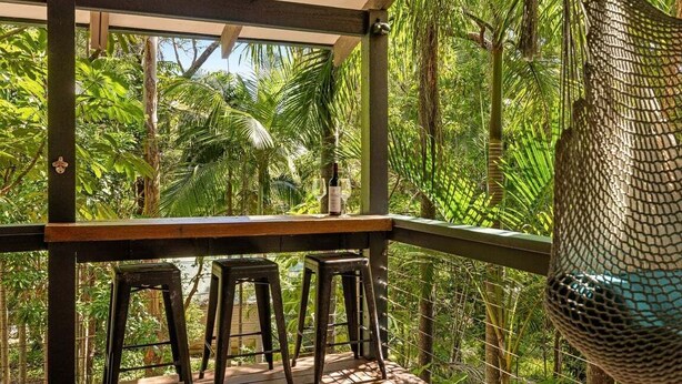 Une terrasse en bois avec une table et deux tabourets, entourée d’une végétation tropicale luxuriante.