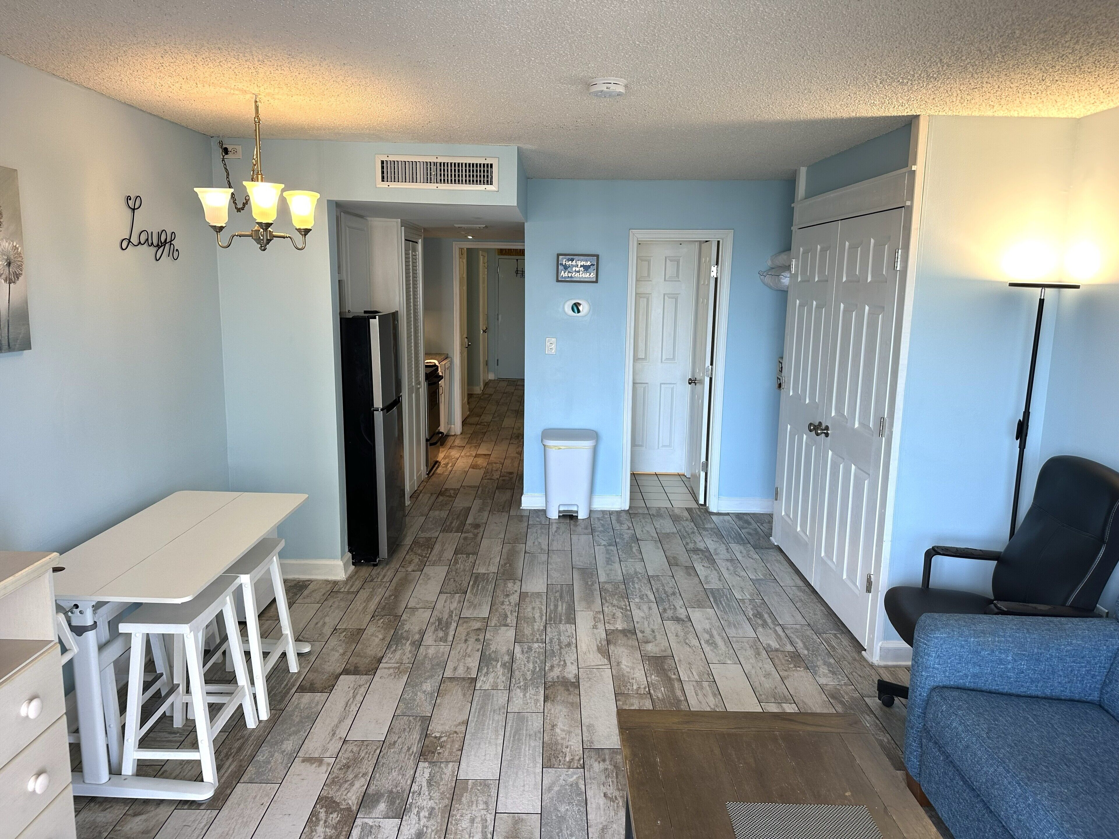 A compact kitchen with a refrigerator, a dining table, and a chair. A hallway leads to a door.