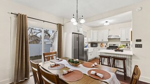 A modern kitchen with white cabinets, a stainless steel refrigerator, and a dining area with wooden chairs and a table set for four. - Haven on Main - 4 Bed 2 Bath (Sioux Falls)