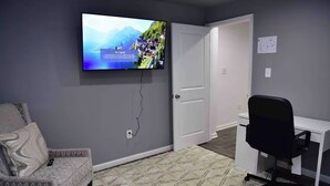 Ett rum med en tv-skärm som visar ett bergigt landskap, en mönstrad matta, en grå soffa och en vit dörr.