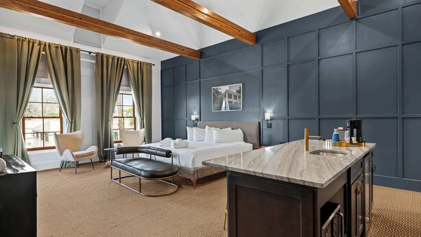 A hotel room with a bed, a desk, a chair, a sofa, a window with curtains, and a dark wall with a picture. - Huge King Suite w. Rain Shower, Balcony, Wet Bar (Beaufort)