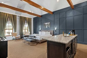 A hotel room with a bed, a desk, a chair, a sofa, a window with curtains, and a dark wall with a picture. - Huge King Suite w. Rain Shower, Balcony, Wet Bar (Beaufort)