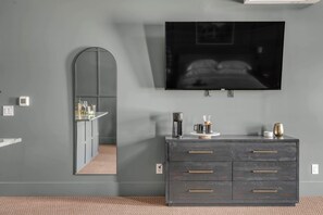 Une chambre moderne avec un mur gris, un grand téléviseur à écran plat fixé au-dessus d’une commode en bois foncé dotée de tiroirs, un miroir et une petite étagère sur laquelle sont posés divers objets.