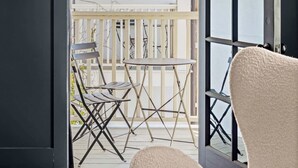 A room with a dark wall, a large window, and a view of a balcony with a chair and a table. - Huge King Suite w. Rain Shower, Balcony, Wet Bar (Beaufort)